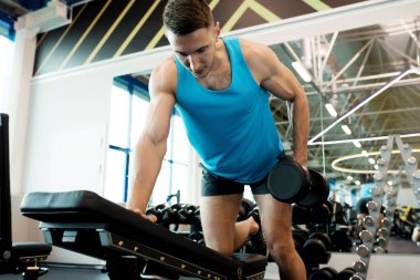 Modern spor salonunda egzersiz sırasında tezgah yaslanmış dambıl ile egzersiz yapan kol kasları pompalama yakışıklı genç adamın düşük açılı portre