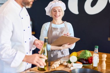 Genç asistanı giyen önlük gerekli not almak süre gülümseyen portresi sakallı ona gösterilen Şef nasıl düzgün yemek hazırlamak için modern restoran mutfak arka plan üzerinde iç