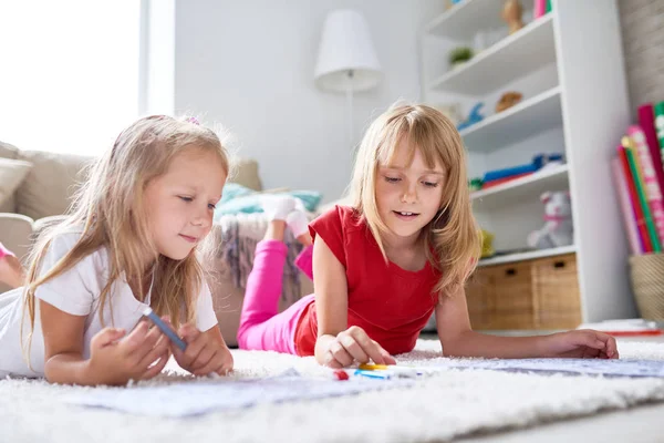 Creative little girls lying on carpet and drawing pictures with felt-tip pens, lovely interior of living room on background