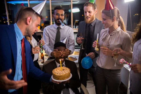 Young office workers congratulating male colleague on birthday, closing his eyes and presenting cake with candle