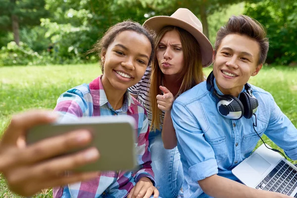 Çok etnik gruptan oluşan grup üç öğrenci selfie açık havada, yeşil çimenlerin üzerine alarak smartphone kameraya görünümlü yapması