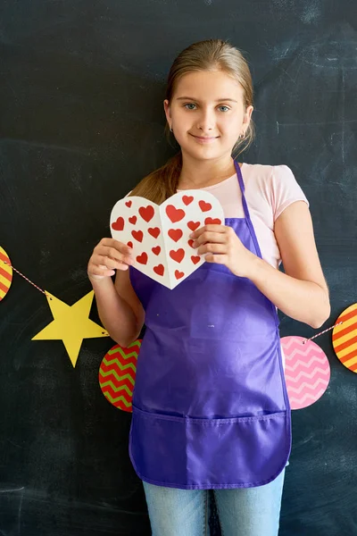 Portrait of  smiling  teenage girl holding paper heart and smiling at camera  posing against blackboard in school