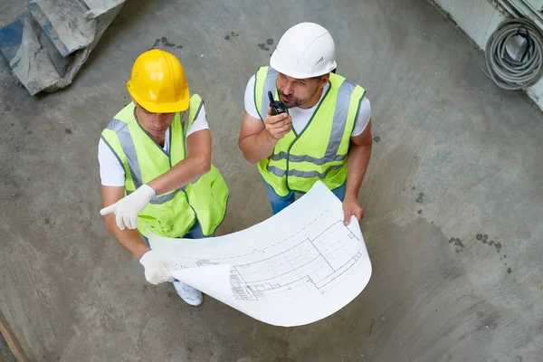 High angle view of busy group of construction workers wearing hardhats and reflective jackets discussing floor plan, bearded middle-aged man talking via walkie-talkie