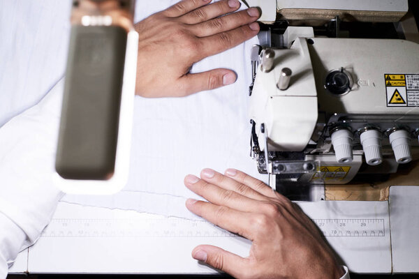 Above view closeup of tailors hands leading white fabric through serger machine working in lamplight at night