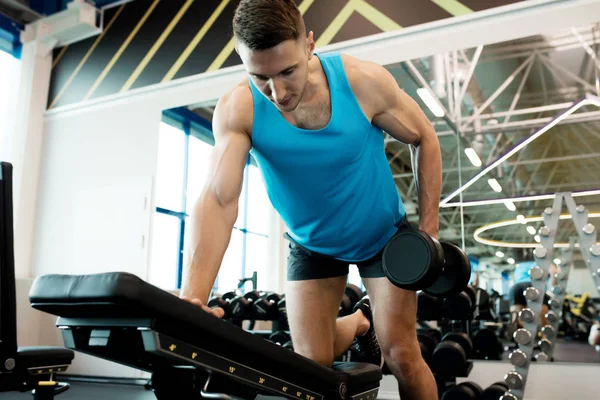 Modern spor salonunda egzersiz sırasında tezgah yaslanmış dambıl ile egzersiz yapan kol kasları pompalama yakışıklı genç adamın düşük açılı portre
