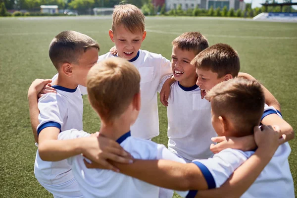 Team of happy boys in uniform embracing during practice on football field