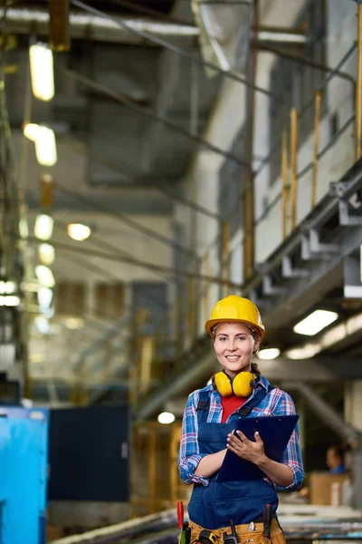 クリップボードを保持しているカメラに微笑んでヘルメットの近代的な工場での作業で 若い女性の肖像画 — ストック写真