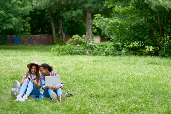 两个女孩坐在绿色草坪上用笔记本电脑的肖像 使用智能手机享受阳光明媚的夏日 — 图库照片