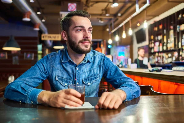 Portrait of drunk young man sitting at table in bar with glass of alcohol looking away pensively and smiling