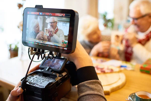 Unrecognizable man recording video of senior couple showing family photos, focus on modern 4k video camera and image on screen