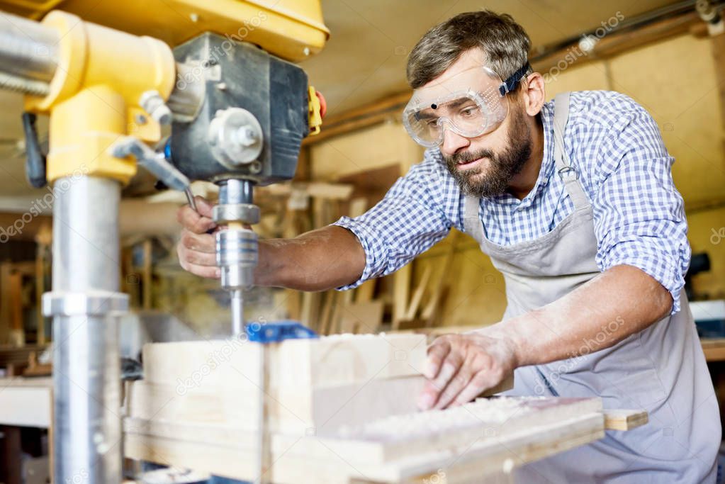 Handsome bearded carpenter wearing safety goggles and apron using drill press machine in order to make holes in wooden plank, interior of spacious workshop on background