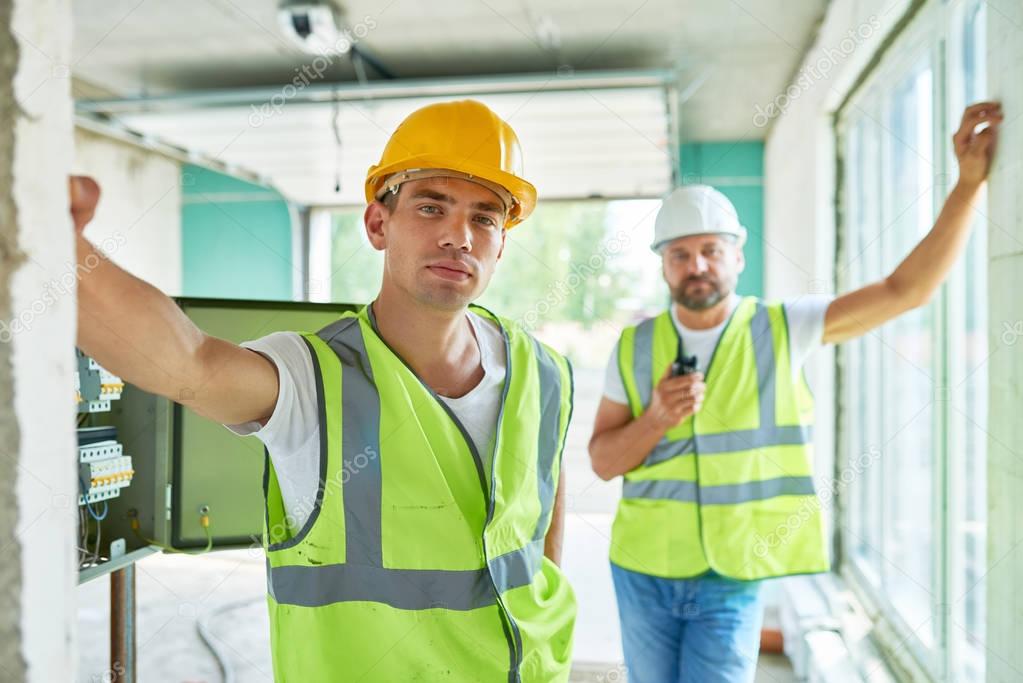 Portrait of two professional builders wearing hardhats and reflective vests  posing confidently looking at camera inside of unfinished building