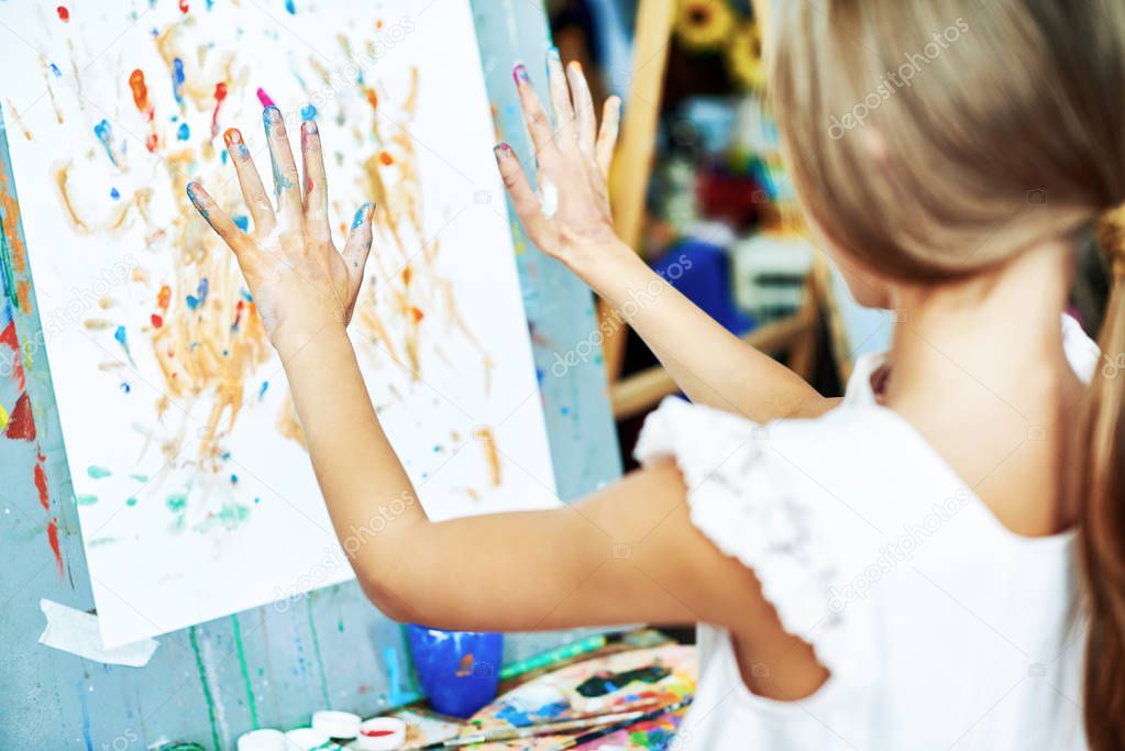 Back view of unrecognizable little girl standing at easel and doing finger-painting, her hands covered with watercolors