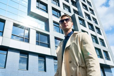 Güneş gözlüğü ve şık trençkot açık havada duran yakışıklı genç işadamı portresi düşük açılı alan kopyalamak uzakta güvenle modern ofis binası, cam önü karşı arıyorsunuz