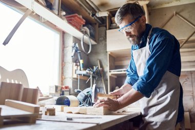 Bearded middle-aged craftsman wearing safety goggles and apron hammering nails into board, interior of messy workshop on background clipart