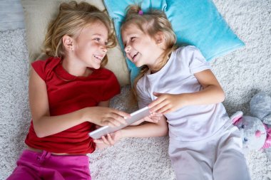 Birbirine bakarak ve katta dijital tablet kullanırken gülümseyen iki mutlu küçük kız portresi görünümü yukarıda evde halı