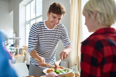 Mutlu bir şekilde kutlama şölen için masada yemek ayarlama arkadaşlarınızla akşam yemeği hazırlarken gülümseyen modern genç adam portresi