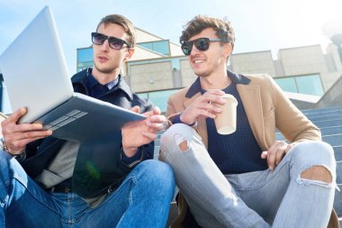 İki yakışıklı genç adam açık havada bina ve kahve keyfini modern Cam merdivenlerde oturan dizüstü kullanarak düşük açılı portresi