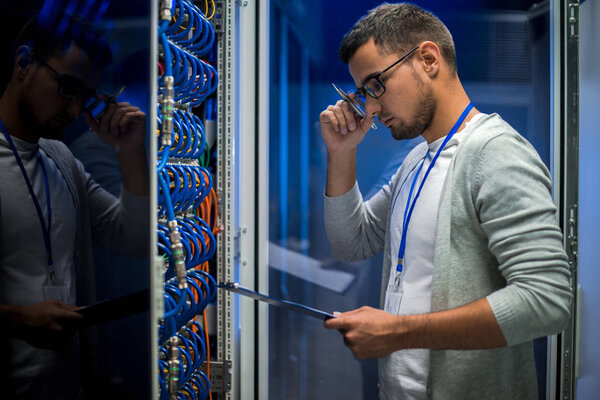 Side view  portrait of young man standing by server cabinet while working with supercomputer in data center and holding clipboard