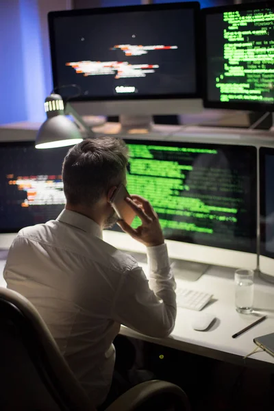 スマート フォン 背景に薄暗いオフィスのインテリアに彼の同僚に話している間現代のコンピューターの前に座って白いシャツを着て認識できないプログラマの背面図 — ストック写真