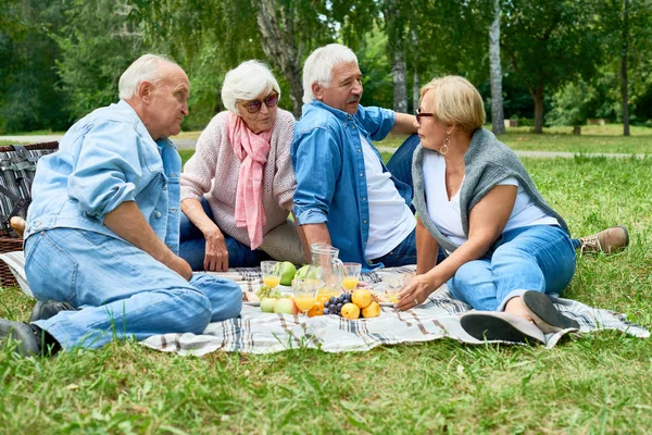 Two senior couples enjoying each others company while having picnic at green park, they sitting on plaid covered with fresh juicy fruits