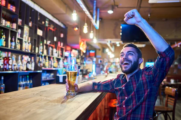 Portrait of excited sports fan cheering happily in pub watching match on TV raising hands and drinking beer, copy space