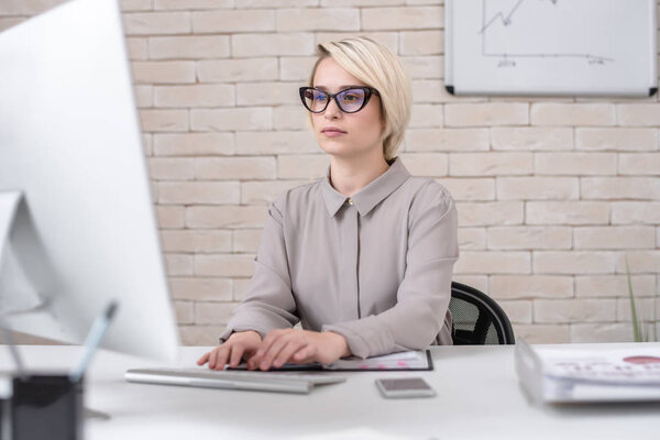 Portrait of blonde successful businesswoman using computer for work sitting at desk in modern office, analyzing data