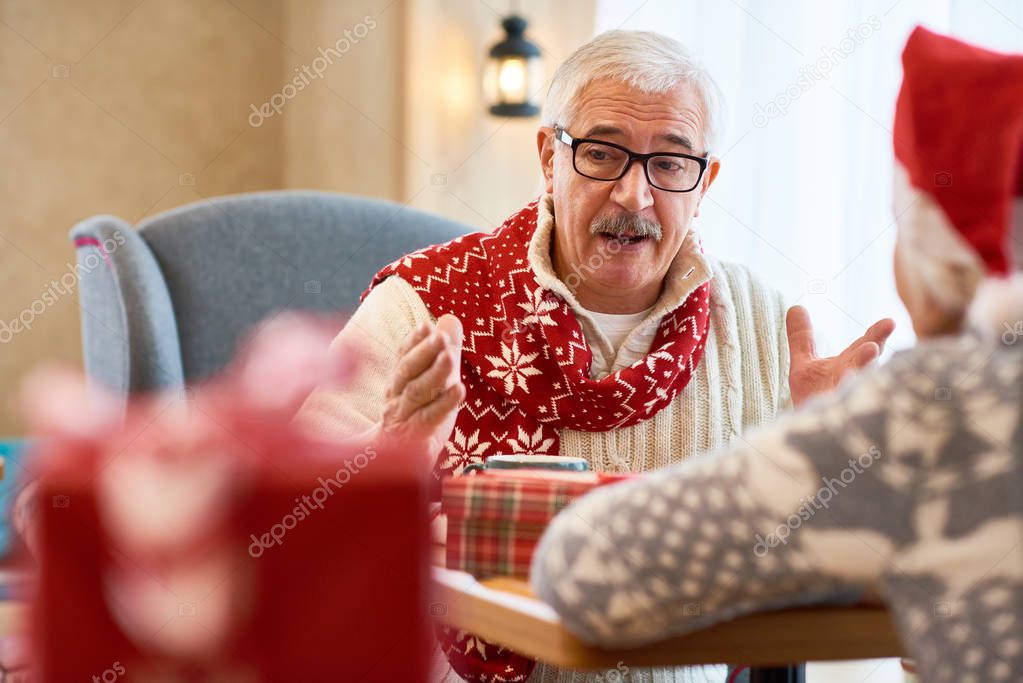 Portrait of white-haired senior man wearing glasses talking to his wife sitting at small table enjoying Christmas holidays at home, copy space