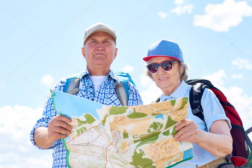 Portrait of active senior couple travelling on hiking trip, stopping to look at map for directions against clear blue sky