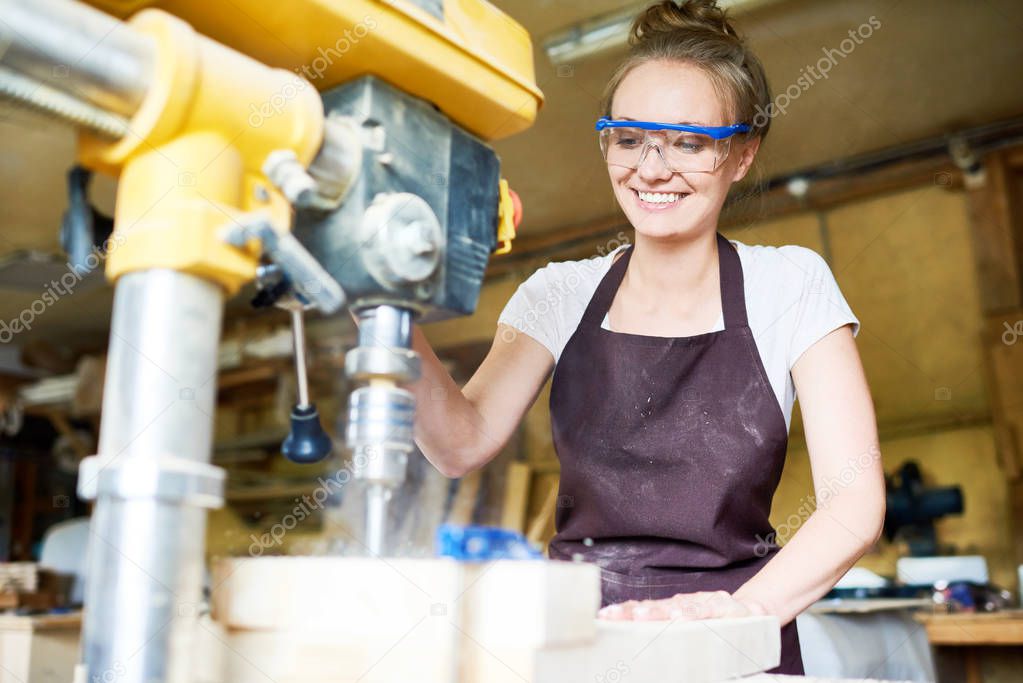 Attractive young woodworker with toothy smile using drill press machine in order to make holes in wooden plank, blurred background