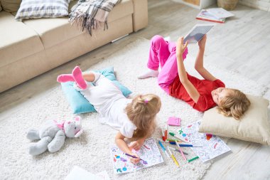 Sarışın küçük kız dijital tablet üzerinde rahat carper, sevimli kız kardeşi Keçeli Kalemler ile resim boyama ve ilgi ile dokunmatik ekran seyir dikkati yatan süre oyun oynama