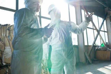 İki işçi biohazard giyen portresi endüstriyel depo modern atık işleme tesisi güneş ışığı penceresinde karşı ayakta uygun