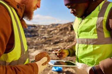 Mid-section portrait of two industrial workers wearing reflective jackets, one of them African, inspecting mineral ore on site outdoors and using digital tablet clipart