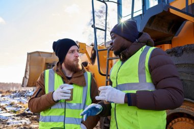 Portrait of two workers, one African-American, drinking coffee and chatting next to heavy industrial truck on worksite clipart