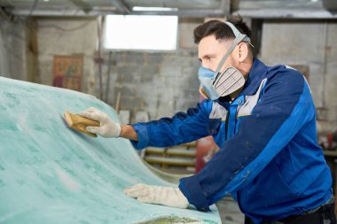 Side view portrait of young  man wearing respirator repairing boat in yacht workshop using polishing tool, copy space clipart