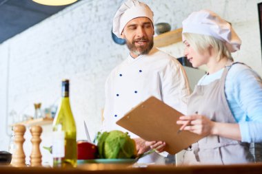 Çanak birlikte, modern restoran mutfak arka plan üzerinde iç hazırlarken oldukça genç asistanı ile Tekdüzen paylaşım mutfak sırlarını giymiş yakışıklı sakallı Şef