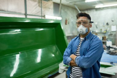 Portrait of handsome workman wearing protective goggles and mask  posing confidently with arms crossed in painting workshop, copy space clipart