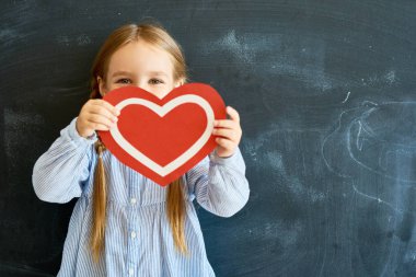 Yüzünü kırmızı kağıt kalp arkasında saklanıyor ve blackboard okulda karşı poz fotoğraf makinesi bakarak sevimli küçük kız portresi