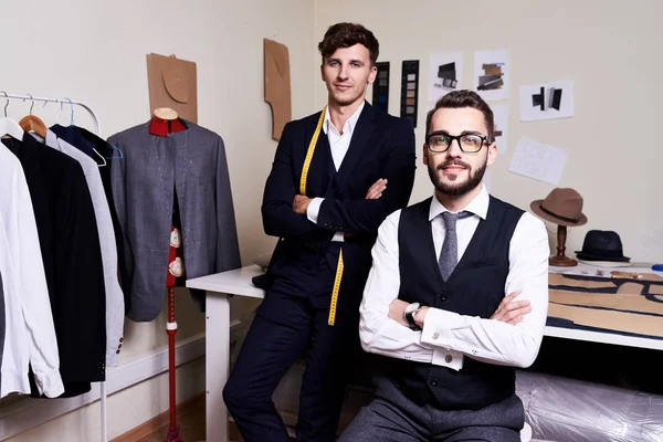 Portrait of two  handsome young tailors posing in traditional atelier studio against working table, looking at camera and smiling confidenty