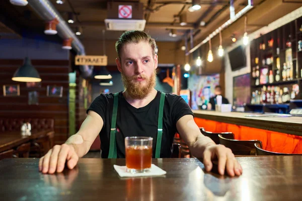 Portrait of modern bearded man getting drunk in pub sitting at table alone and drinking whiskey