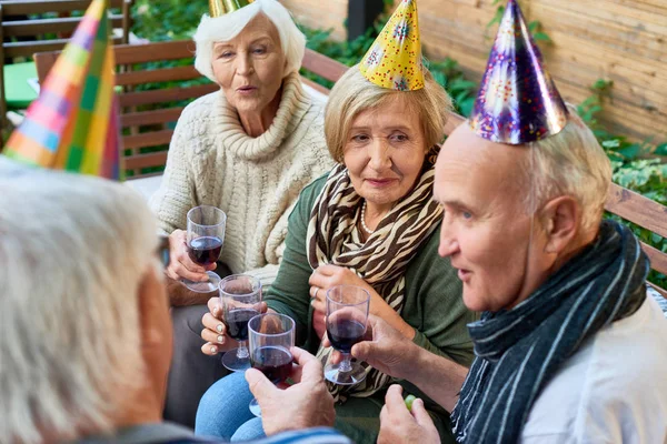 Group of elderly friends wearing warm clothes holding glasses with red whine in hands and chatting with each other while having outdoor birthday party