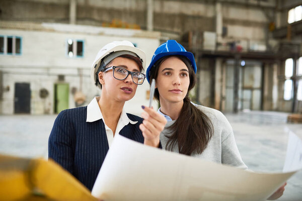 Группа инженеров-технологов в защитных шлемах проводит неформальную встречу в производственном отделе современного завода: обсуждают свои идеи и изучают чертежи
