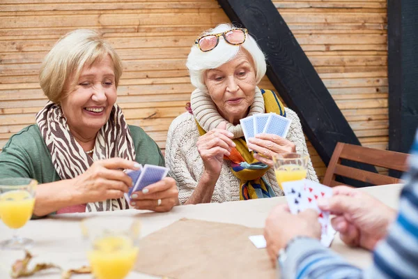 Joyful senior women wearing warm clothes playing poker with friends while sitting at outdoor cafe table