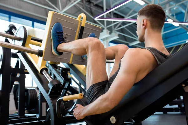 Rear view of muscular sportsman doing legs exercise using machine in modern gym
