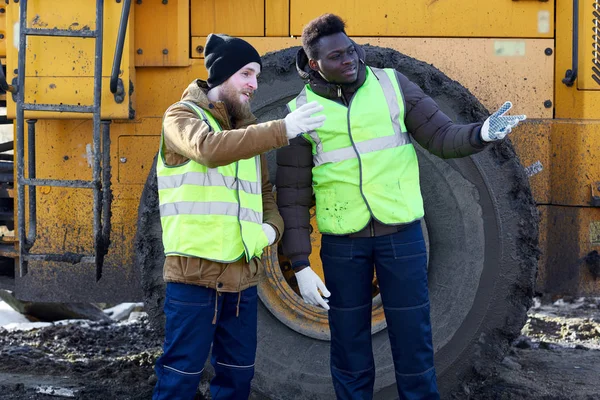 Portrait of two workers, one African-American, standing in dirt next to heavy industrial dump truck on worksite