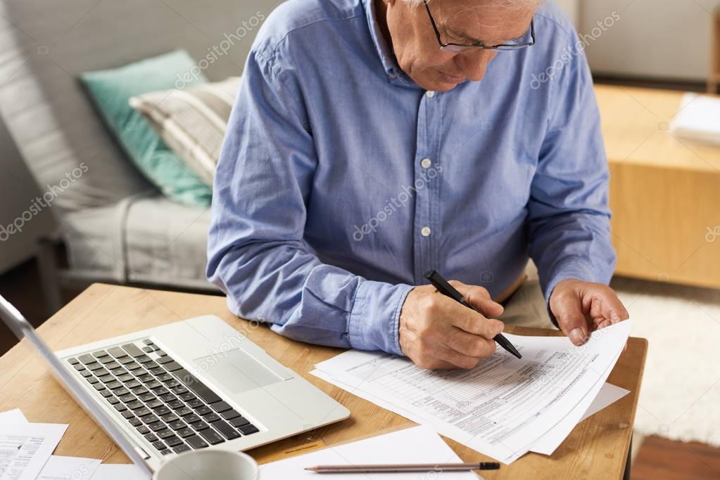 Portrait of modern senior man filling in application form  while working with laptop at home in cozy living room
