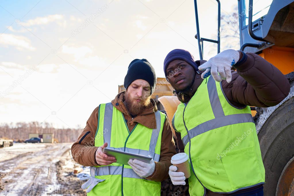 Portrait of two workers, one African-American, drinking coffee and using digital tablet standing next to heavy industrial truck on worksite, copy space