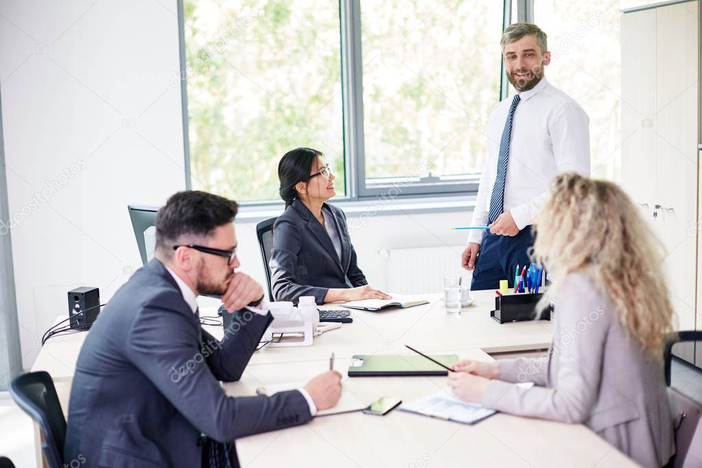 Working process at modern boardroom: handsome bearded manager presenting his ideas while standing at marker board, his colleagues sitting at table and listening to him with interest