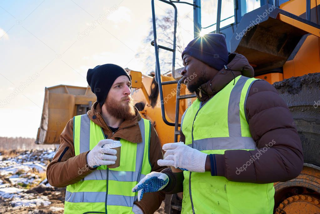 Portrait of two workers, one African-American, drinking coffee and chatting next to heavy industrial truck on worksite