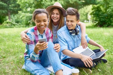 Yeşil çimenlerin üzerine oturmuş ve yaz günü üzerinde mola zevk akıllı telefon ekranına bakarak gülümseyen üç mutlu öğrenci portresi